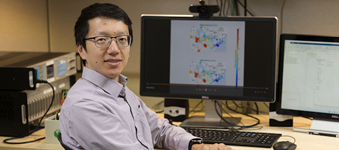 Professor Ziang Zhang wearing glasses seated at a desk in a research lab, with computer monitors displaying scientific data and diagrams