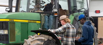 Individuals inspect and take notes on a John Deere tractor inside an agricultural workshop