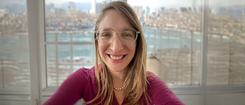 Professional headshot of a person wearing glasses, a necklace, and a magenta long-sleeve top, photographed indoors with large windows overlooking a waterfront and distant city skyline