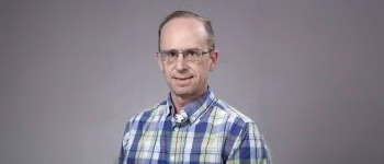 Studio portrait of a person wearing glasses and a blue, white, and green plaid button-down shirt, facing the camera against a plain gray background