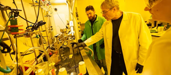 Three researchers wearing lab coats, safety glasses, and protective gloves work around specialized equipment in a laboratory. One researcher points toward an instrument on a crowded laboratory bench while two others observe. Tubing, glassware, gauges, and containers are visible throughout the workspace, which is illuminated by bright yellow lighting commonly used in nanotechnology or semiconductor