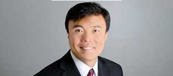 Professional headshot of professor Jun Qu from University at Buffalo wearing a dark suit jacket, white dress shirt, and red patterned tie against a plain light gray background