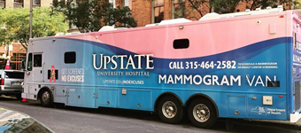 A large mobile mammography van from Upstate University Hospital is parked along a city street. The vehicle features a blue and pink exterior with prominent text promoting breast cancer screening services, including a phone number to schedule a mammogram. Buildings, trees, and parked vehicles line the street in the background