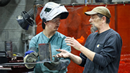 A student wearing a welding helmet, safety glasses, protective sleeves, and heavy gloves holds a fabricated metal piece while receiving instruction from an instructor in a welding lab. The instructor gestures toward the project as the two discuss the work. Welding equipment and a red protective welding curtain are visible in the background