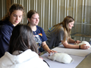Students participate in a hands-on animal science activity, gathered around a table with two white rabbits. One student gently handles a rabbit while others observe and interact with the animals. The setting appears to be an agricultural or veterinary learning space, with wire enclosures and metal-paneled walls visible in the background