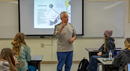 A presenter stands at the front of a classroom-style training session, speaking to seated attendees. A presentation slide is displayed on a projection screen behind the presenter, while participants sit at tables facing the front of the room. The classroom has white walls, bright lighting, and a professional learning environment, with notebooks, laptops, and other materials visible on the tables