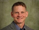 Professional headshot of a person wearing a gray blazer and blue collared shirt against a mottled green studio background