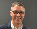 Professional headshot of a person wearing glasses, a dark jacket, and a white collared shirt against a gray background