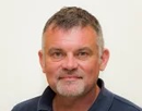 Professional headshot of a person with short gray hair wearing a dark collared shirt against a light background