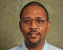Professional headshot of professor Dali Ismail wearing round eyeglasses and a light blue collared shirt against a muted green studio background