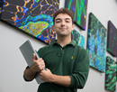 P3 PharmD student Francis Depalo in a green shirt holding a laptop in a hallway filled with paintings