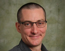 Professional headshot of associate professor Julien Panetier from Binghamton University wearing rectangular glasses and a dark patterned button-down shirt up against a mottled green studio background