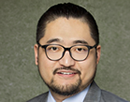 Professional headshot of assistant professor Kaicheng Yang wearing round black-framed glasses, a light blue dress shirt, and a gray suit jacket, photographed against a neutral textured background