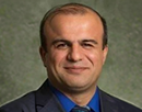 Professional headshot of a person wearing a dark suit jacket, blue dress shirt, and matching patterned tie against a neutral, mottled gray-green studio background
