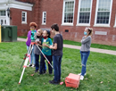 Students use a surveying instrument on a tripod during an outdoor learning activity beside a brick school building.