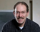 Professional headshot of distinguished professor Tim Lowenstein wearing glasses and a dark plaid button-down shirt against an indoor background that includes part of a chalkboard