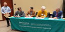 Panel discussion featuring several seated speakers behind a table with a university-branded banner, while another speaker stands at the front of the room