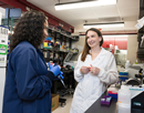Two researchers in lab coats work together in a laboratory, discussing samples and equipment at a workstation with test tubes, pipettes, and scientific instruments