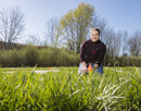 Assistant professor Amber Churchill kneeling in a grassy field with tall vegetation, conducting outdoor agricultural or environmental research. Trees and shrubs are visible in the background under a cloudy sky