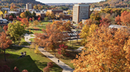 A wide aerial view of a university campus in autumn, with academic buildings surrounded by trees displaying vibrant red, orange, and yellow foliage. Walking paths wind through green spaces, and a few people are visible crossing the campus