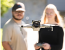 A drone hovers in the foreground with its camera facing forward, while two people stand blurred in the background outdoors. One person holds a tablet or controller used to operate the drone. The scene is set in daylight with a softly focused natural backdrop, emphasizing drone technology and remote operation