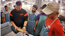 Students and an instructor wearing protective aprons gather around a fish tank in an aquaculture facility as the instructor holds a fish and demonstrates handling techniques. Rows of tanks and water system equipment are visible in the background