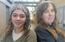 Two individuals stand side by side in an academic building hallway, posing for a portrait with classrooms and display cases visible in the background