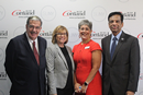 Four people in professional attire pose in front of a SUNY Cortland step-and-repeat backdrop displaying the university logo and website. The group stands side by side for a formal event photo, with two people wearing business suits, one wearing a light-colored blazer, and one wearing a bright red dress. The backdrop features repeated SUNY Cortland branding on a white background