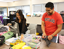 Two students organize backpacks, school supplies, and activity materials on a table in a classroom or office setting. Storage bins filled with colorful items and stacks of supplies are visible nearby