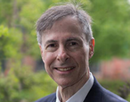Professional headshot of a person wearing a dark suit jacket, white dress shirt, and light blue patterned tie. The person is facing the camera with a slight smile. The background is softly blurred with green foliage and hints of an outdoor setting, creating a professional portrait appearance