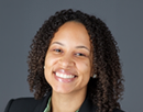 Professional headshot of a person with curly hair wearing a dark blazer against a gray studio background