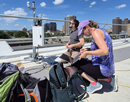 Two people work with monitoring equipment on a rooftop overlooking a city skyline on a bright day with scattered clouds. One person kneels beside an open laptop connected to air-quality monitoring instruments, while another stands nearby next to the mounted sensors and data collection equipment