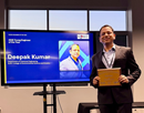 A person stands beside a presentation screen while holding a framed award or plaque. The screen displays a slide featuring the person's photo and name, indicating recognition for an achievement or honor