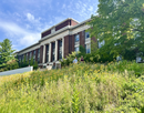 Historic brick academic building with large white columns overlooks a hillside filled with wildflowers and native grasses, with two people walking along a path in front of the building