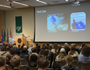 Presenter speaks to a large audience in an auditorium while displaying slides about animal tool use on a projection screen. An ESF banner and flags are visible beside the stage