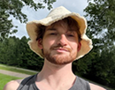 Close-up outdoor portrait of a person wearing a light-colored bucket hat and a gray sleeveless shirt. The person is standing near a paved path surrounded by green trees. Sunlight illuminates the face and hat, while a partly cloudy sky is visible overhead