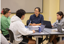 Four people sit around a table in a classroom or meeting room engaged in a small-group discussion. Papers and materials are spread across the table as participants collaborate