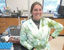 Associate professor Jennifer Gilbert-Jenkins in a laboratory wearing a white lab coat, standing beside a workbench with scientific equipment and sample trays