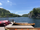 Wooden docks extend into a calm lake on a sunny day. A row of red kayaks is stored along the shoreline to the left, while forested hills and rocky outcrops surround the water in the distance. The sky is clear blue with a few wispy clouds, creating a scenic lakeside view