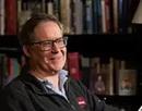 Professor Matthew Hendley wearing glasses and a dark pullover sits in a chair holding papers, pictured in front of bookshelves filled with books in a library or office setting with warm lighting