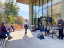 People gather outside a building to watch a live music performance, with musicians singing and playing instruments near the entrance and audience members seated on benches and colorful chairs