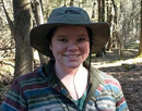 Associate professor Mary Alldred wearing a wide-brimmed hat and striped outdoor shirt stands in a wooded area, surrounded by trees and forest vegetation