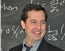 Professional headshot of a person wearing a dark jacket over a collared shirt and tie, standing in front of a chalkboard. The chalkboard behind the person contains handwritten mathematical equations and symbols written in white chalk. The image is closely cropped around the head and shoulders, with the chalkboard providing an academic or classroom setting in the background
