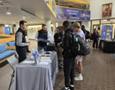 Several attendees stand and talk at an information table inside a large event hall. The table, covered with a white cloth, displays handouts and promotional materials. In the background, additional tables, banners, and framed displays line the room, while groups of attendees mingle and network along a carpeted walkway under bright indoor lighting