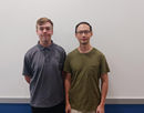Two researchers stand side by side in front of a blank presentation screen in a classroom or meeting space. One wears a gray polo shirt and the other a green T-shirt