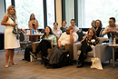 A group of adults sits in a semicircle in a modern meeting space while one participant stands at the front speaking. Large windows line the room, and attendees appear engaged in a discussion or workshop