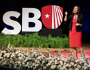 Stony Brook University President Andrea Goldsmith on stage beside large “SBU” letters and logo delivering her second State of the University Address at the Staller Center on September 14, with floral arrangements in the foreground