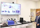 A person in business attire stands in a laboratory or research workspace, looking toward a large wall-mounted display screen showing multiple project dashboards, charts, and data visualizations. Computer monitors, electronic equipment, and workstations are arranged beneath the screen, highlighting a technology-focused environment used for research, monitoring, or project management
