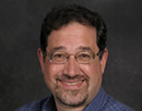 Professional headshot of distinguished professor Daniel Klein wearing wire-rimmed glasses and a blue checkered button-down shirt against a dark gray studio background