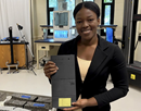 Darla-Ena Boyer holding a model of a “pendulum wall” in a laboratory holding a black test sample, standing beside construction material specimens and research equipment. Laboratory instruments and workstations are visible in the background